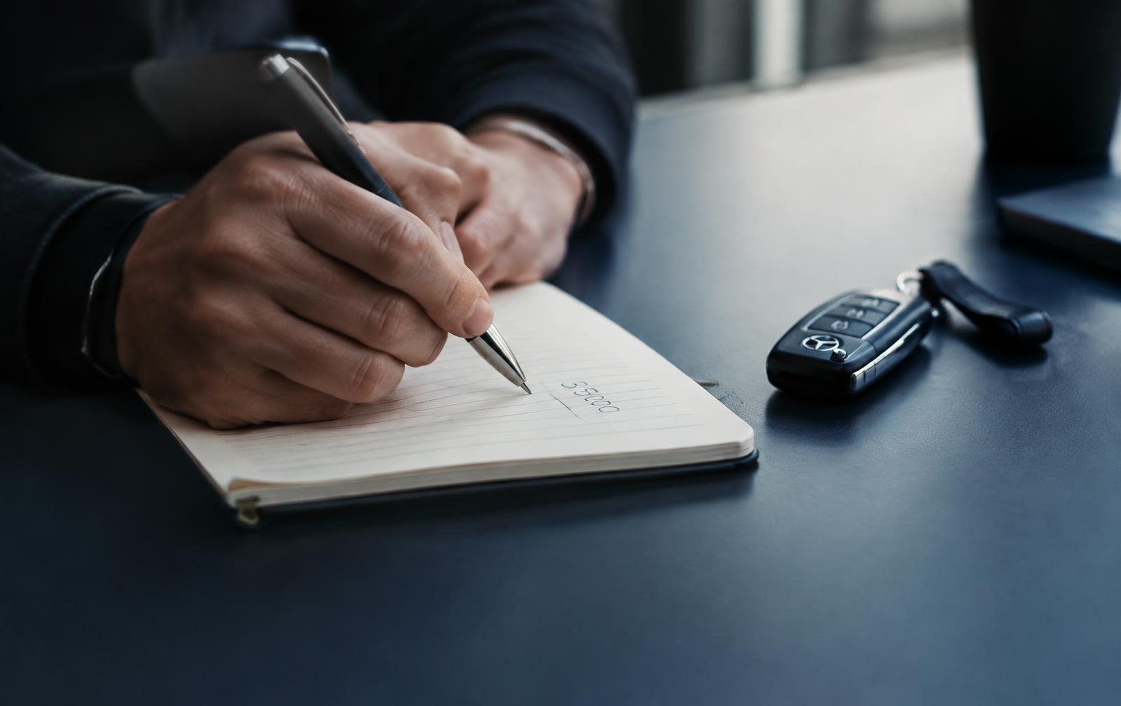A person writing a price figure in a notebook beside car keys